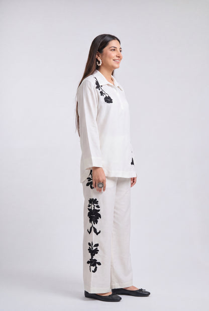 A woman modeling a white two-piece kurta set with black floral embroidery, standing in profile against a light gray studio background.