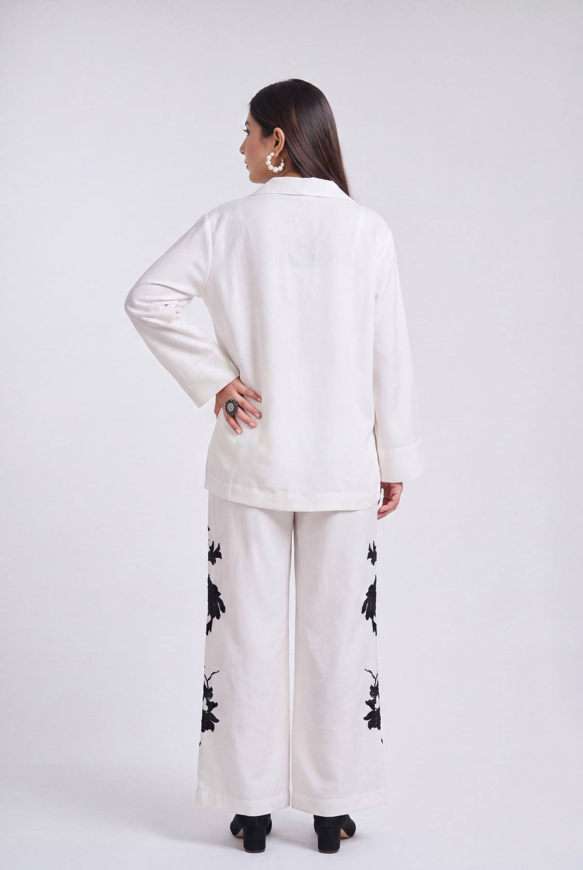 A woman shown from the back wearing a white long-sleeve top and matching wide-leg pants with black floral embroidery, photographed against a light gray studio background.