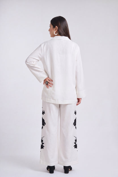 A woman shown from the back wearing a white long-sleeve top and matching wide-leg pants with black floral embroidery, photographed against a light gray studio background.