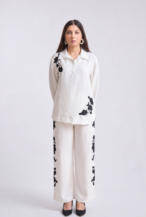 A woman modeling a matching two-piece outfit consisting of a long-sleeve collared top and wide-leg trousers in off-white, both featuring black floral embroidery, set against a plain light gray studio background.