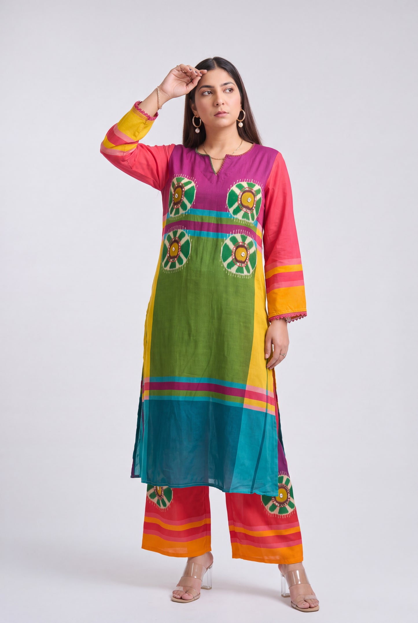 A woman modeling a vibrant, multicolored traditional Indian kurta and trouser set. The outfit features a rainbow-like vertical stripe pattern with green, yellow, blue, pink, and orange panels, along with circular floral motifs on the chest and matching striped pants.