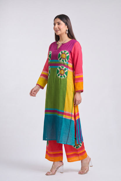 A woman modeling a vibrant, multicolored traditional Indian kurta and trouser set against a plain light gray studio background. The outfit features a rainbow-like color-blocked pattern with green, yellow, pink, orange, and blue panels, along with embroidered floral motifs on the chest.