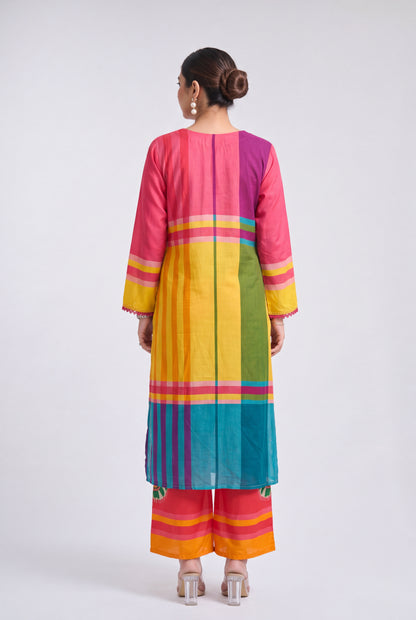 A woman viewed from the back wearing a brightly colored striped kurta and matching wide-leg pants. The outfit features vertical panels of pink, purple, yellow, green, blue, orange, and red with thin horizontal accent stripes, set against a plain light gray studio background.