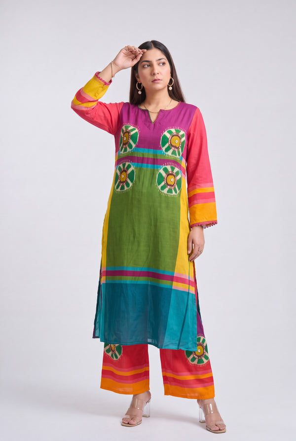 A woman modeling a vibrant, multicolored traditional Indian kurta and trouser set. The outfit features a rainbow-like vertical stripe pattern with green, yellow, blue, pink, and orange panels, along with circular floral motifs on the chest and matching striped pants.