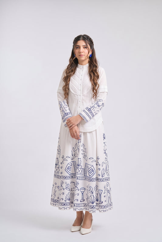 A woman modeling a long white embroidered dress with blue traditional patterns, standing against a light gray studio background.