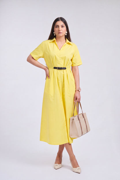 A woman modeling a bright yellow short-sleeve midi shirt dress with a black belt, holding a beige tote bag and wearing beige heels, against a light gray studio background.