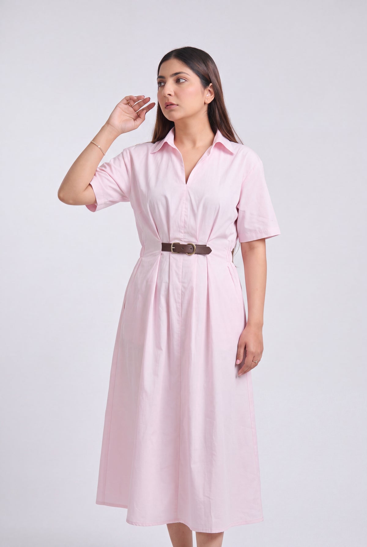 A woman modeling a light pink short-sleeve collared midi dress with a brown belt, standing against a plain light gray studio background.