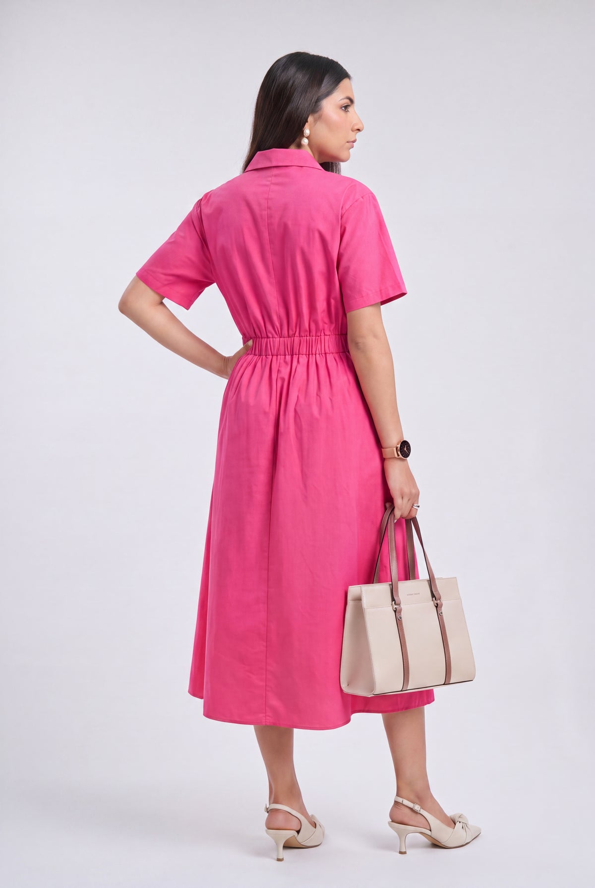 A woman wearing a bright pink short-sleeve midi shirt dress, shown from the back. She is holding a beige tote bag and wearing beige high-heeled sandals, set against a plain light gray studio background.