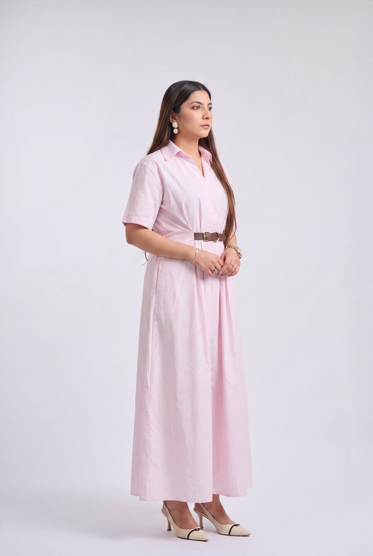 A woman modeling a light pink short-sleeve collared maxi dress with a matching belt, standing against a plain light gray studio background.