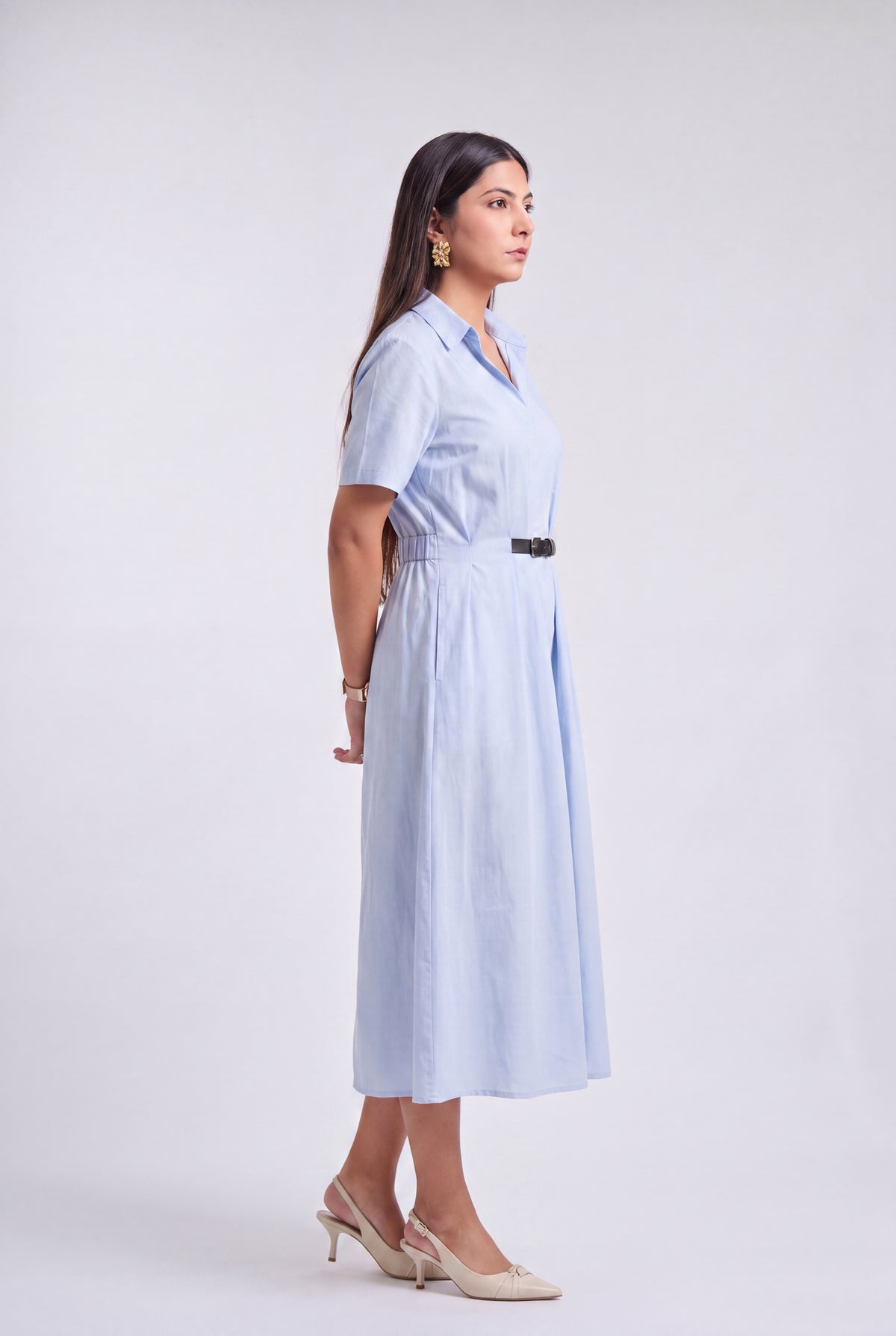 A woman modeling a light blue midi shirt dress with short sleeves, a collared neckline, and a thin black belt at the waist, photographed against a plain light gray studio background.