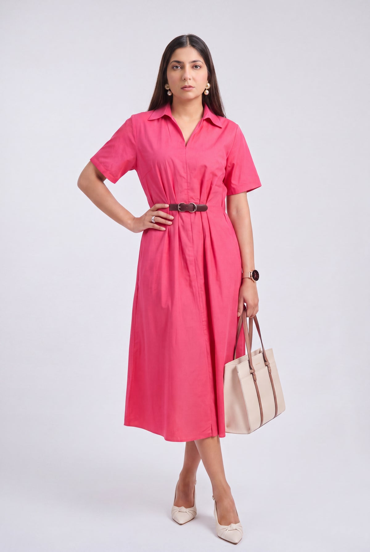 A woman models a short-sleeve pink midi shirt dress with a black belt, holding a beige tote bag and wearing beige heels, against a light gray studio background.