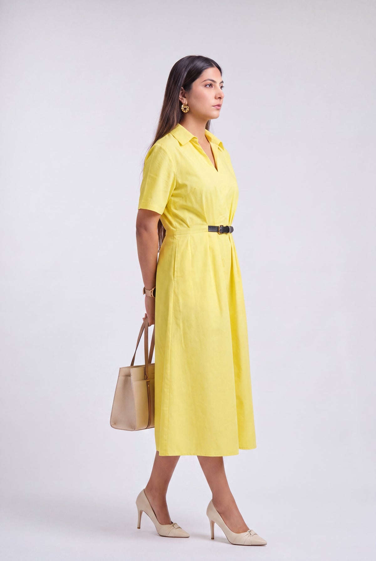 A woman modeling a bright yellow short-sleeve midi dress with a black belt, styled with beige heels and a beige handbag against a light gray studio background.