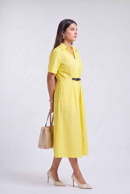 A woman modeling a bright yellow short-sleeve midi dress with a black belt, styled with beige heels and a beige handbag against a light gray studio background.