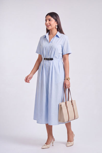 A woman modeling a light blue short-sleeve midi shirt dress with a black belt, holding a beige tote bag and wearing beige heels, against a light gray studio background.
