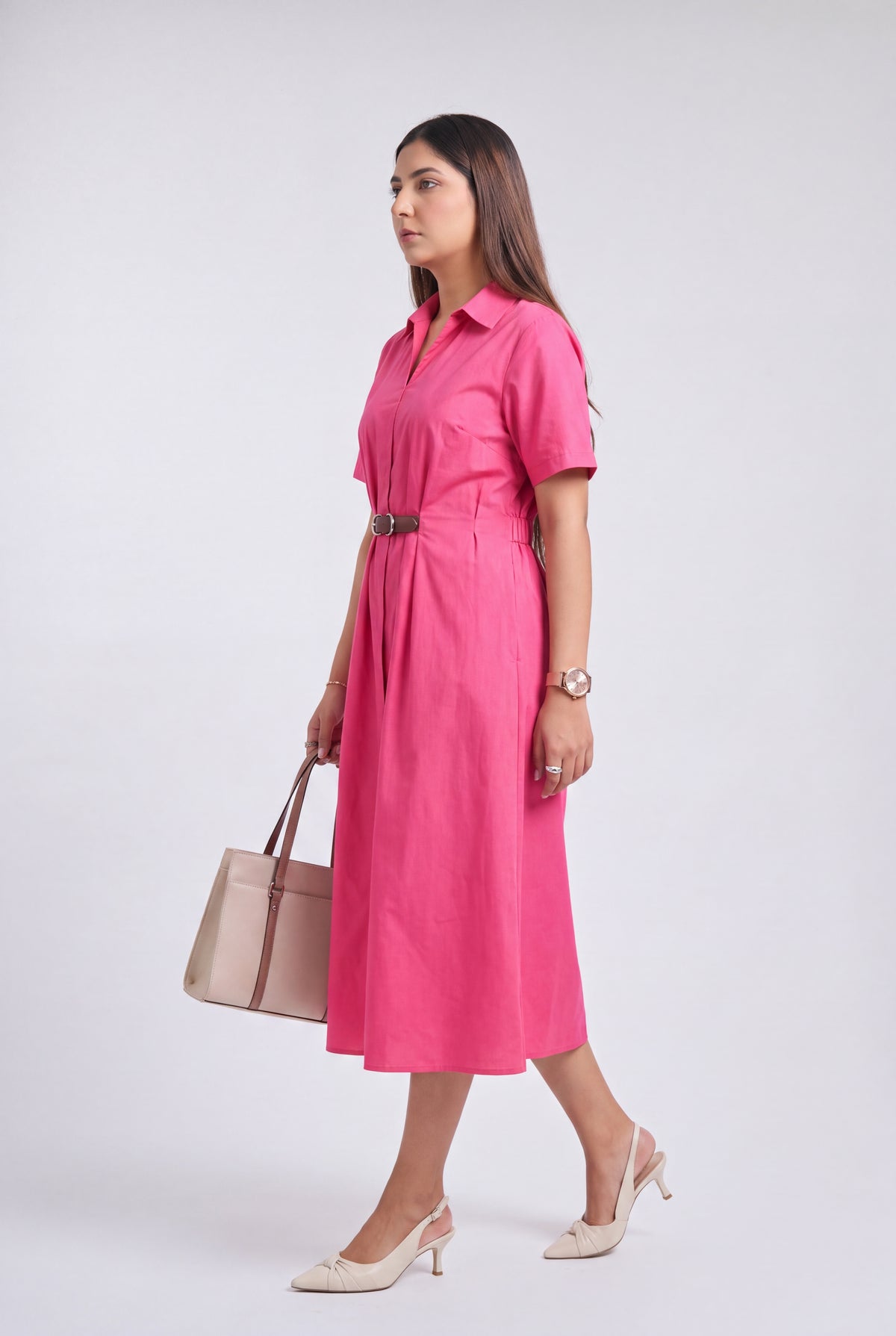 A woman modeling a bright pink short-sleeve midi shirt dress with a matching belt, holding a beige tote bag and wearing beige heels, set against a plain light gray studio background.