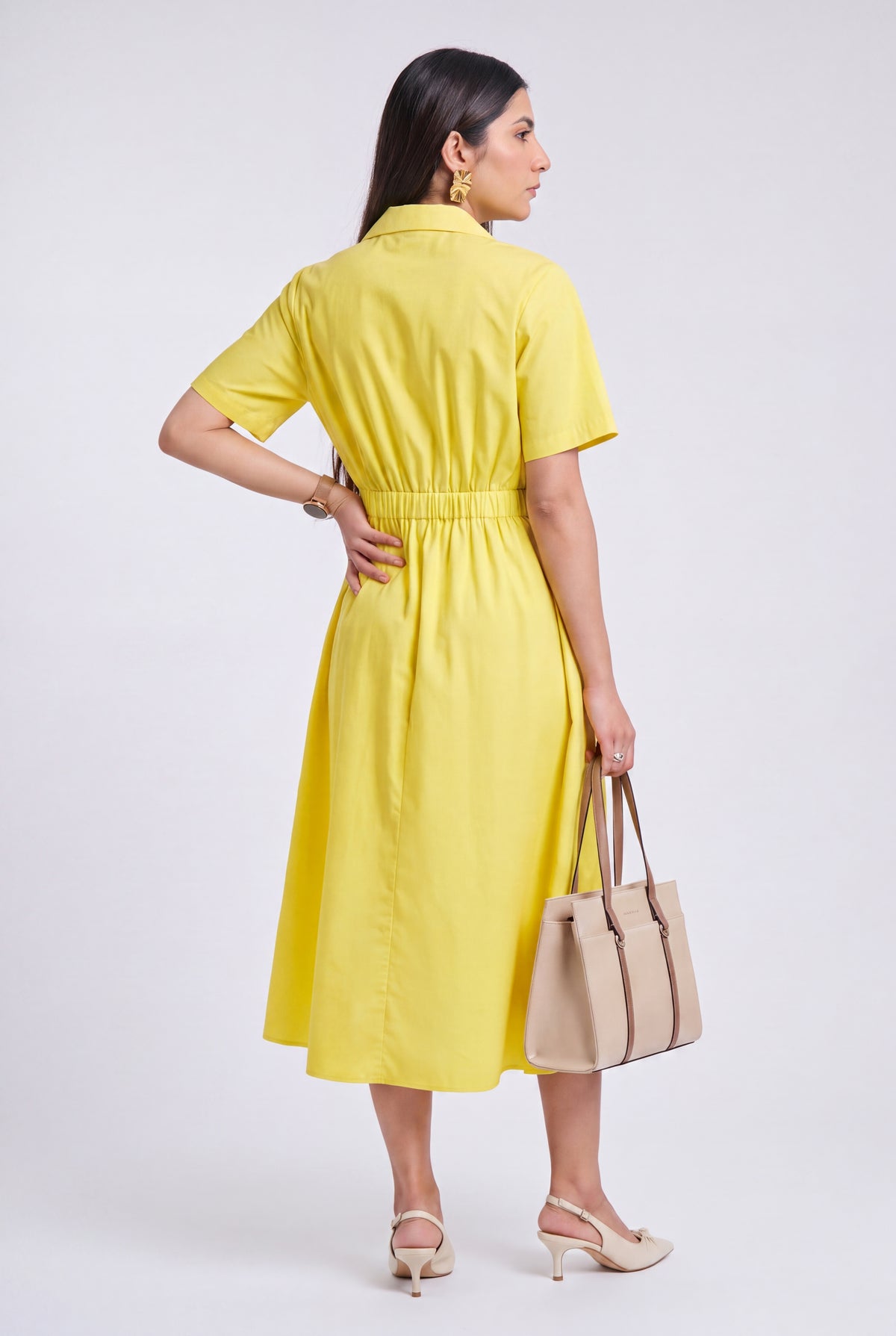 A woman modeling a bright yellow short-sleeve midi dress from the back, holding a beige tote bag and wearing beige heels against a light gray studio background.