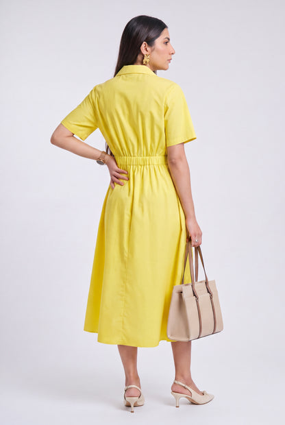 A woman modeling a bright yellow short-sleeve midi dress from the back, holding a beige tote bag and wearing beige heels against a light gray studio background.