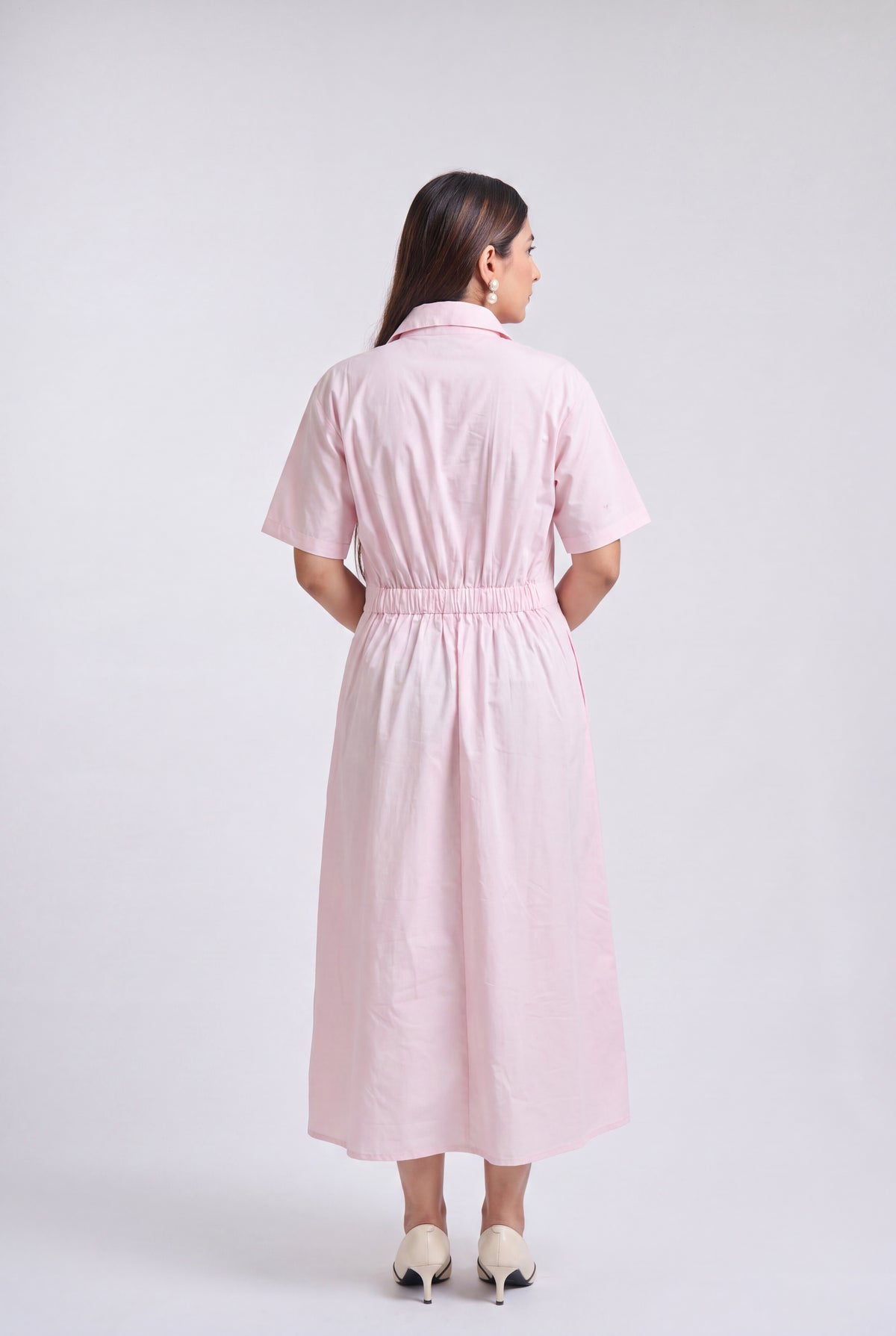 A woman is shown from the back wearing a light pink short-sleeve midi dress with a button-up collar and a gathered elastic waist, paired with beige high heels, against a light gray studio background.