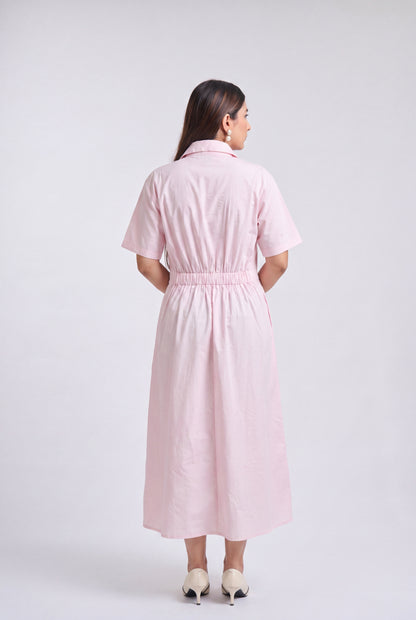 A woman is shown from the back wearing a light pink short-sleeve midi dress with a button-up collar and a gathered elastic waist, paired with beige high heels, against a light gray studio background.