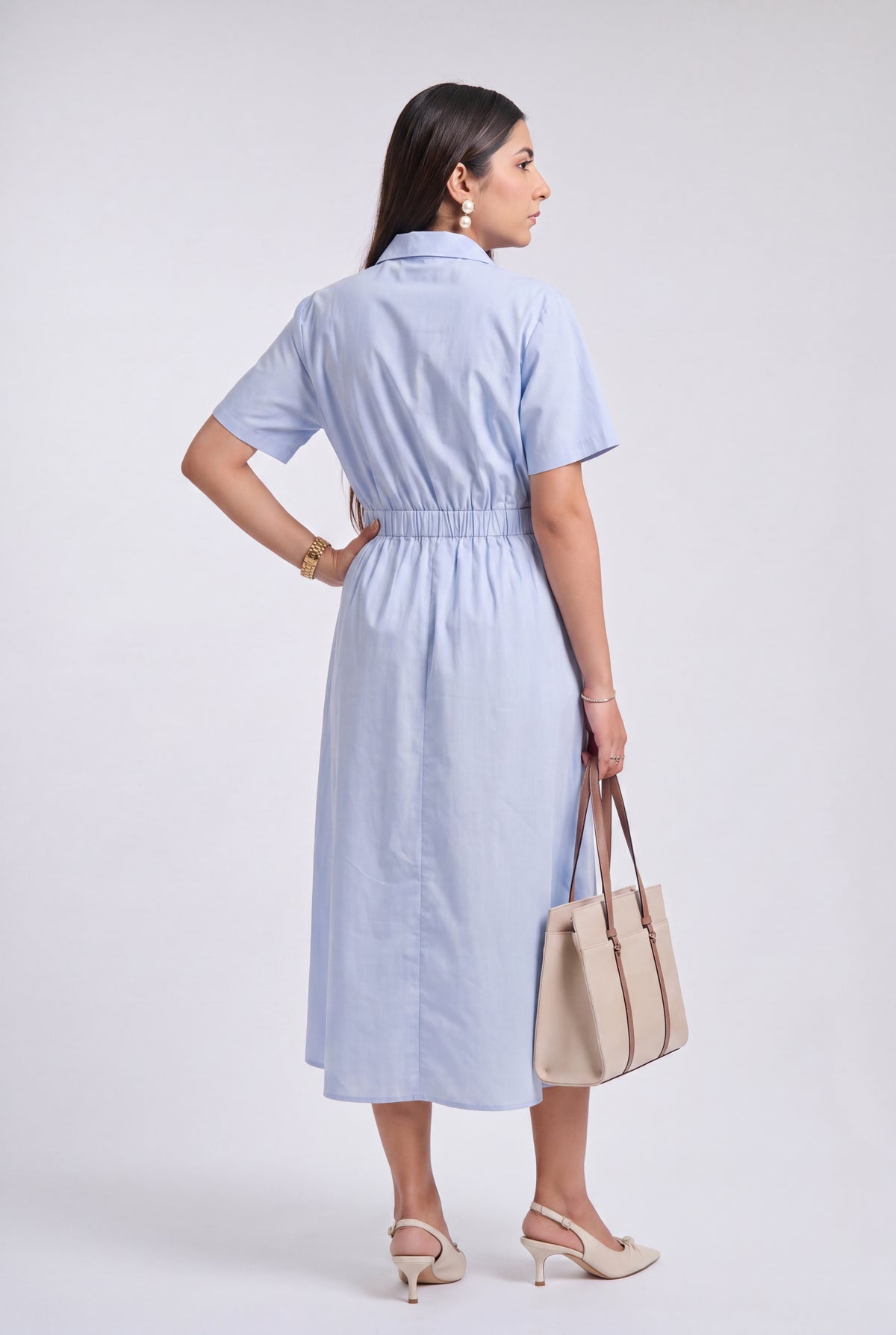 A woman modeling a light blue short-sleeve midi shirt dress from the back, holding a beige tote bag and wearing beige heels against a light gray studio background.