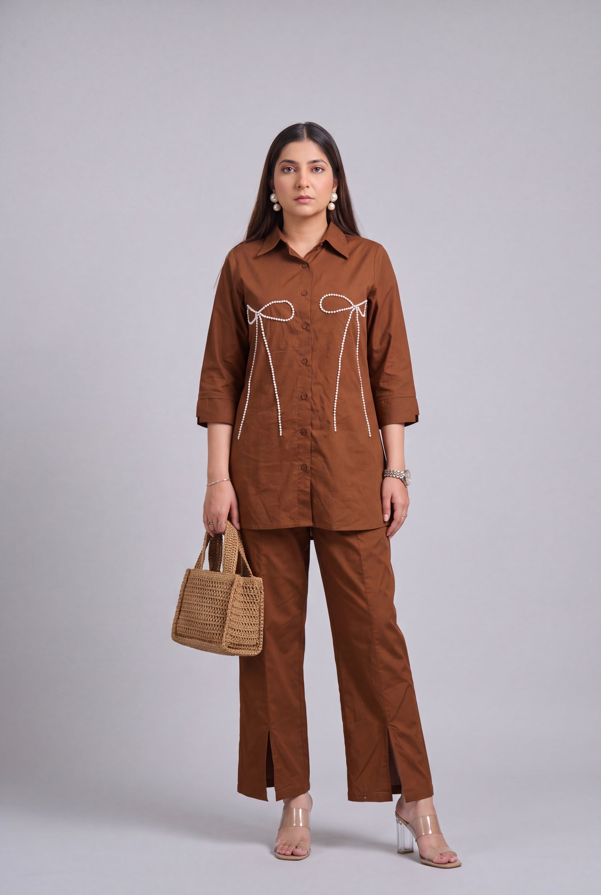 A woman models a matching brown two-piece set consisting of a long-sleeve button-up shirt with white tie details on the chest and matching wide-leg trousers. She is holding a small woven tan handbag and wearing clear high-heeled sandals against a solid gray studio background.