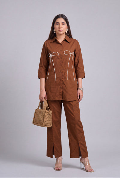 A woman models a matching brown two-piece set consisting of a long-sleeve button-up shirt with white tie details on the chest and matching wide-leg trousers. She is holding a small woven tan handbag and wearing clear high-heeled sandals against a solid gray studio background.