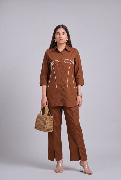 A woman models a matching brown two-piece set consisting of a long-sleeve button-up shirt with white tie details on the chest and matching wide-leg trousers. She is holding a small woven tan handbag and wearing clear high-heeled sandals against a solid gray studio background.