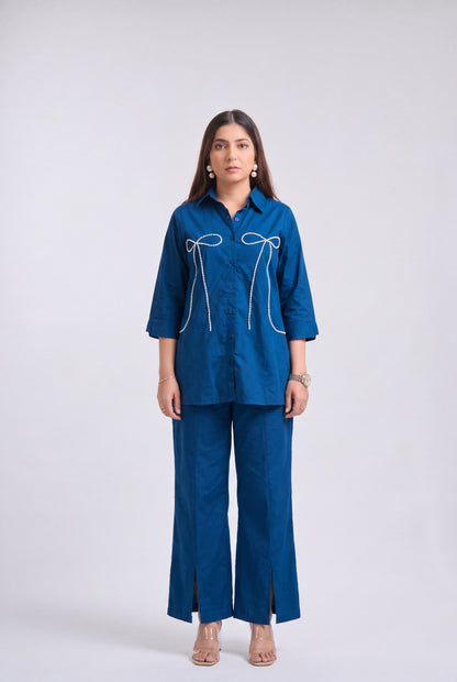 A woman modeling a matching blue two-piece set consisting of a long-sleeve button-up shirt with white bow embroidery and matching wide-leg trousers, shot against a light gray studio background.