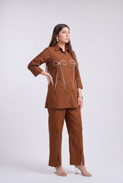 A woman modeling a brown two-piece linen pajama set consisting of a long-sleeve button-up top with white cord details and matching wide-leg pants, shot against a light gray studio background.