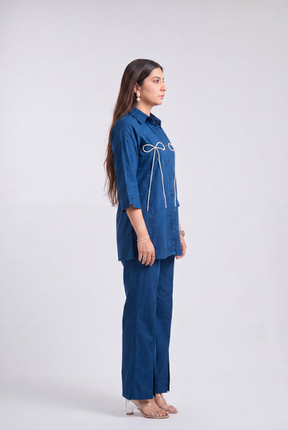 A woman stands in profile wearing a blue matching two-piece outfit with a collared long-sleeve top featuring white embroidered bow details and matching wide-leg trousers, photographed against a light gray studio background.