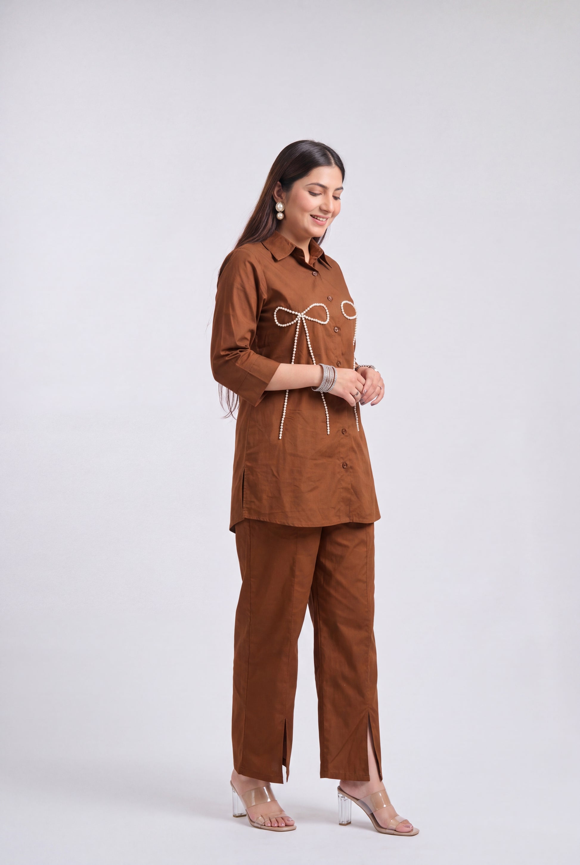 A woman modeling a matching brown two-piece outfit consisting of a long-sleeve button-up shirt with a white drawstring detail at the chest and matching brown trousers, set against a plain light gray studio background.