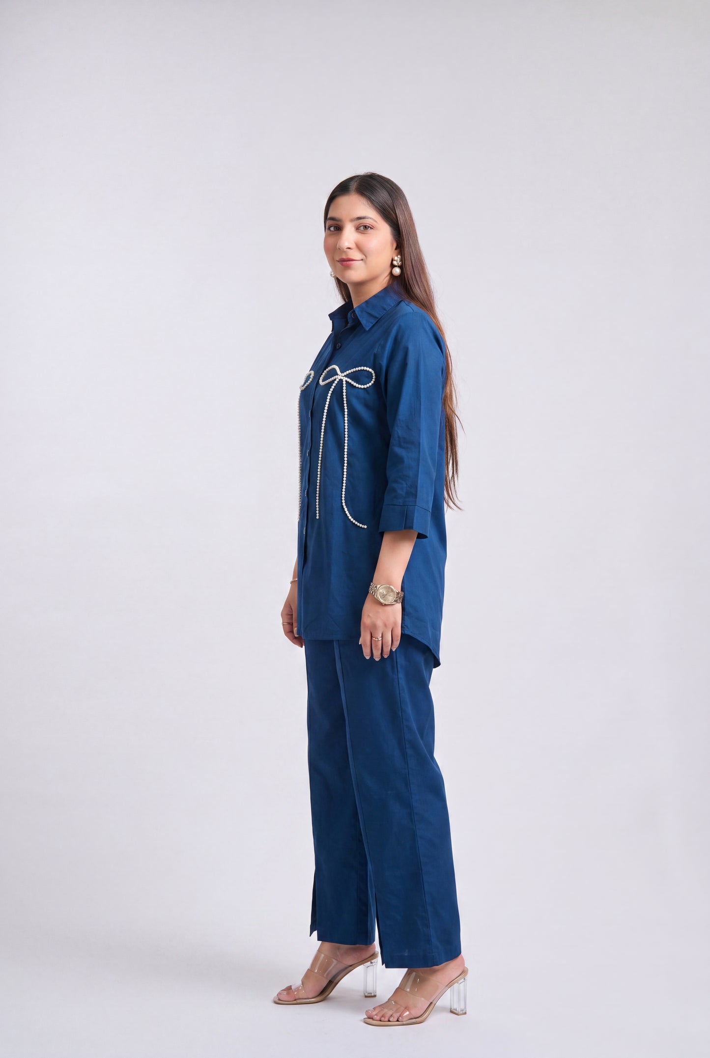 A woman modeling a blue two-piece outfit consisting of a collared long-sleeve top with white decorative trim and matching wide-leg trousers, standing against a light gray studio background.