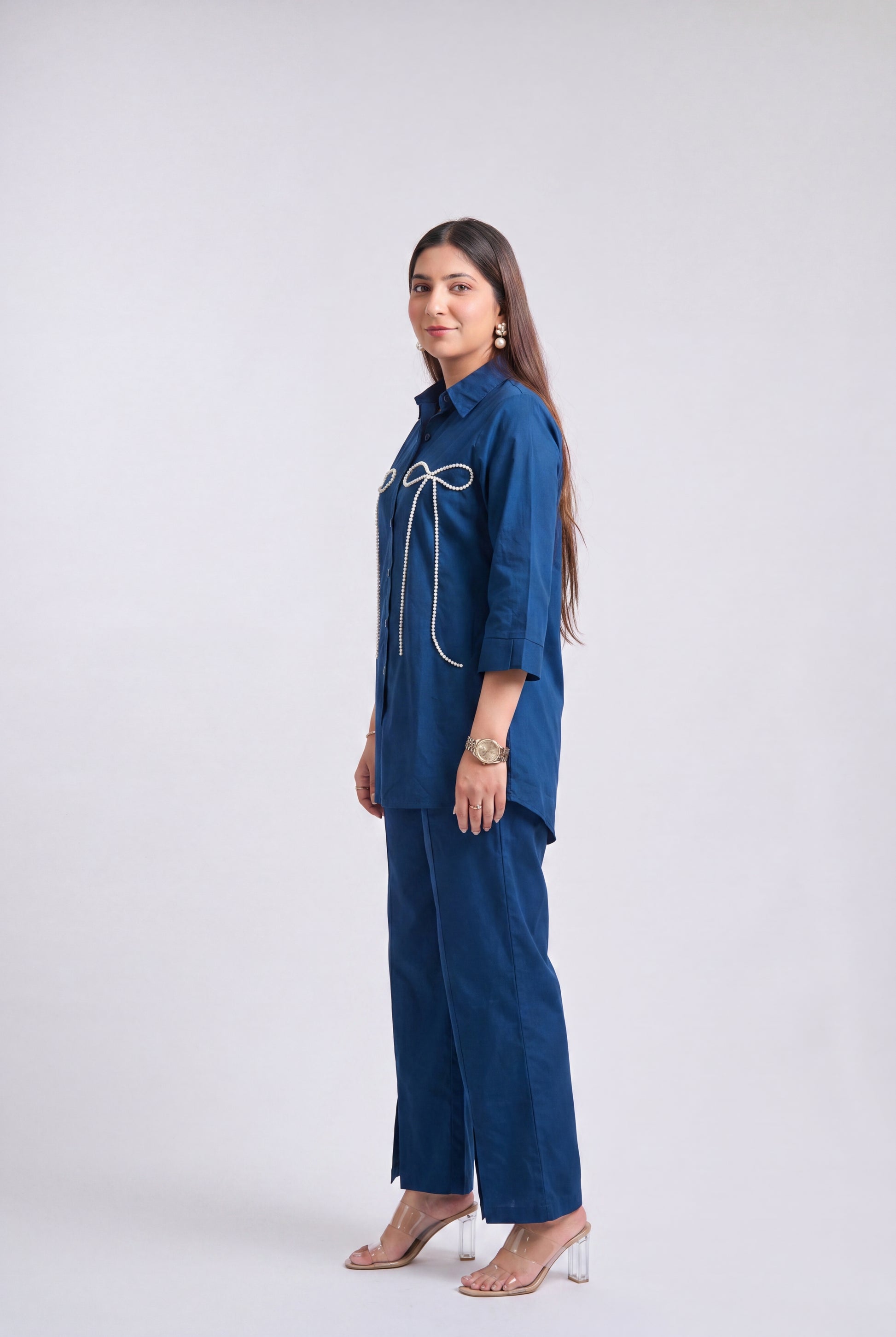 A woman modeling a blue two-piece outfit consisting of a collared long-sleeve top with white decorative trim and matching wide-leg trousers, standing against a light gray studio background.