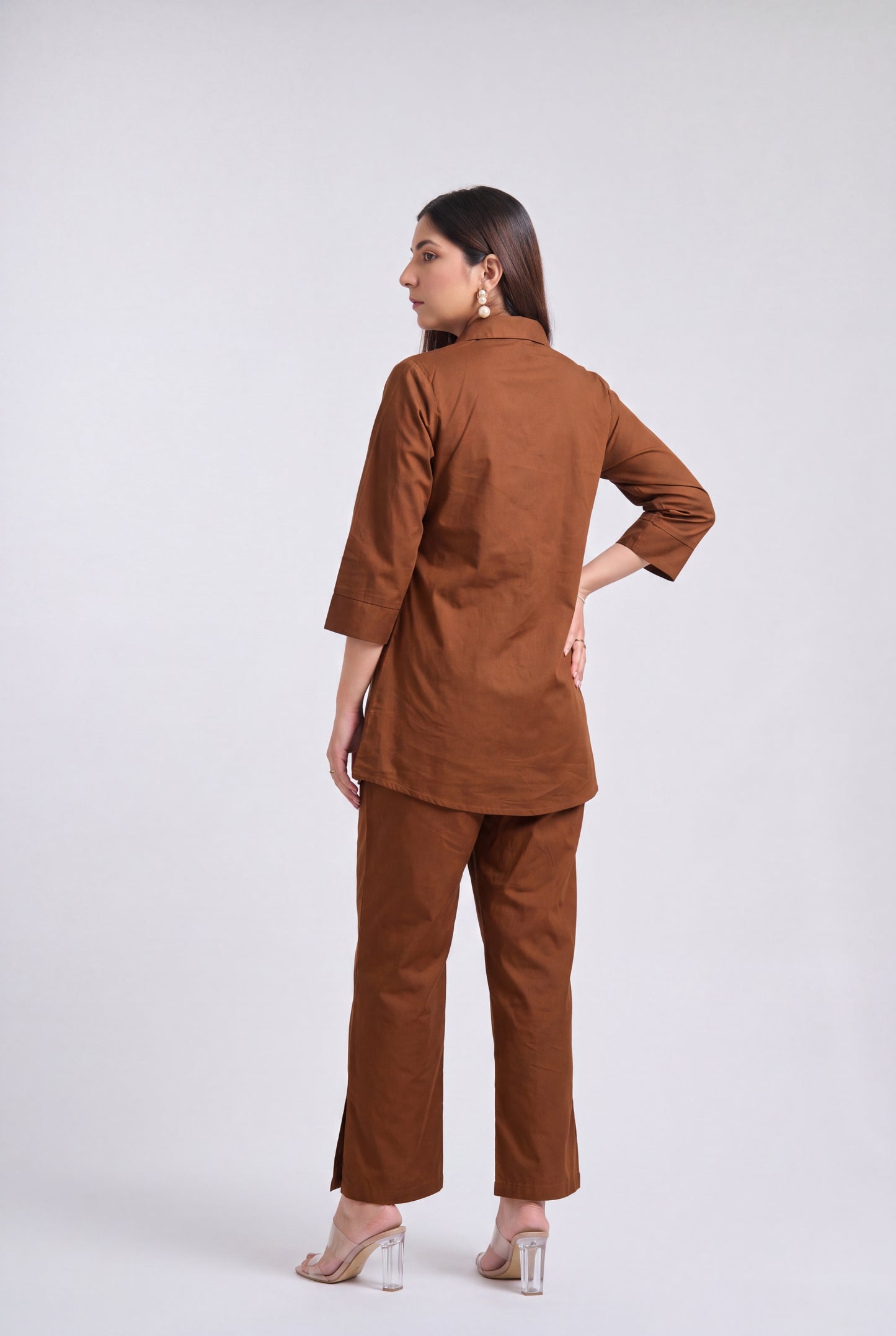 A woman modeling a matching brown two-piece kurta and trouser set, shown from a rear three-quarter angle against a light gray studio background.