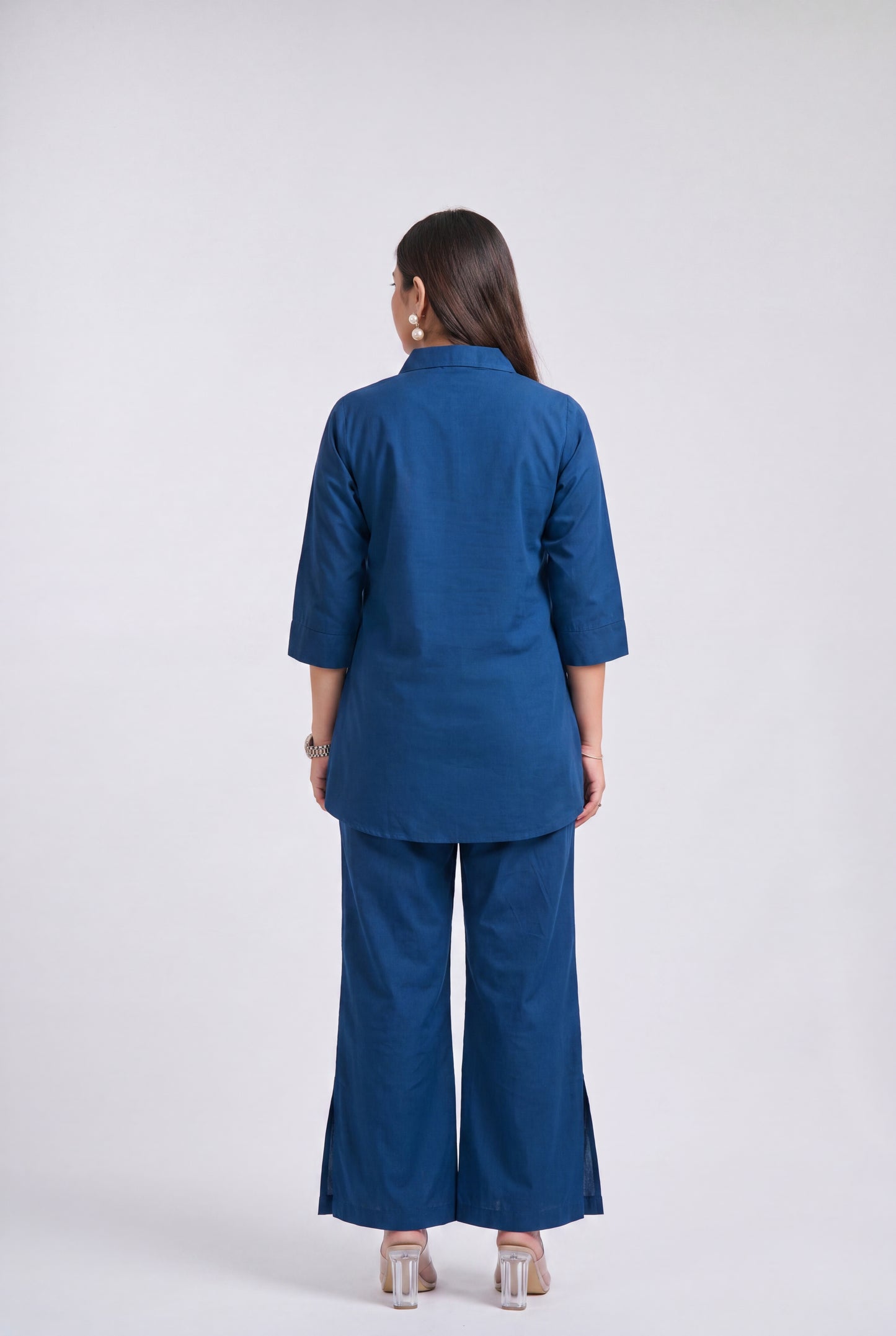 A woman viewed from the back wearing a matching blue two-piece outfit consisting of a short-sleeve collared top and wide-leg pants, paired with clear high-heeled sandals against a light gray studio background.