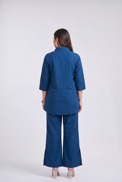 A woman viewed from the back wearing a matching blue two-piece outfit consisting of a short-sleeve collared top and wide-leg pants, paired with clear high-heeled sandals against a light gray studio background.