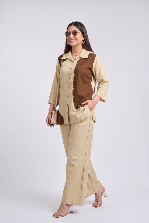 A woman modeling a beige and brown color-blocked two-piece set consisting of a long-sleeve button-up shirt and matching wide-leg trousers, styled with clear high-heeled sandals and a small handbag against a light gray studio background.