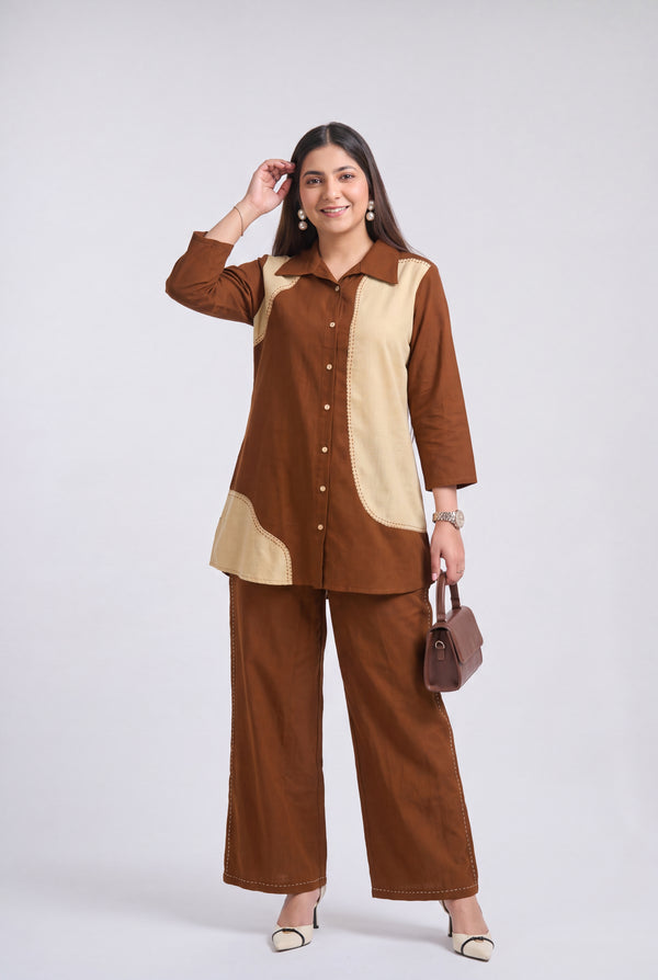 A woman modeling a two-piece brown and beige color-blocked kurta set, featuring a long-sleeve button-up top and matching wide-leg trousers, paired with a small brown handbag and white heels against a light gray background.
