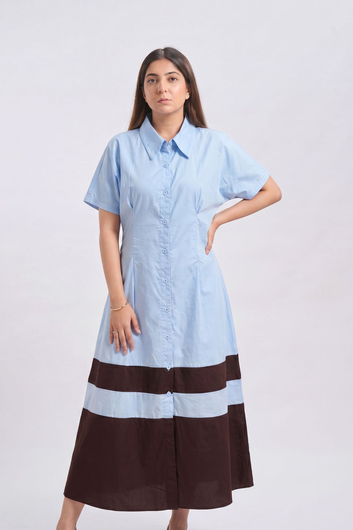 A woman modeling a light blue short-sleeve button-up shirt dress with a dark brown and light blue striped hem, set against a plain light gray studio background.