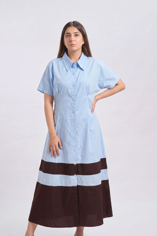 A woman modeling a light blue short-sleeve button-up shirt dress with a dark brown and light blue striped hem, set against a plain light gray studio background.