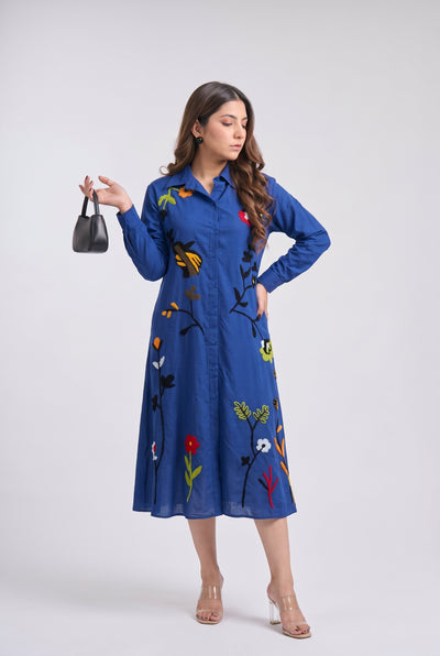 A woman modeling a blue long-sleeve midi shirt dress with colorful floral and butterfly embroidery, standing against a light gray studio background.