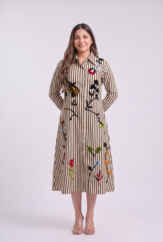 A woman modeling a long-sleeve midi shirt dress with vertical beige and brown stripes and colorful embroidered floral and butterfly motifs, set against a plain light gray studio background.