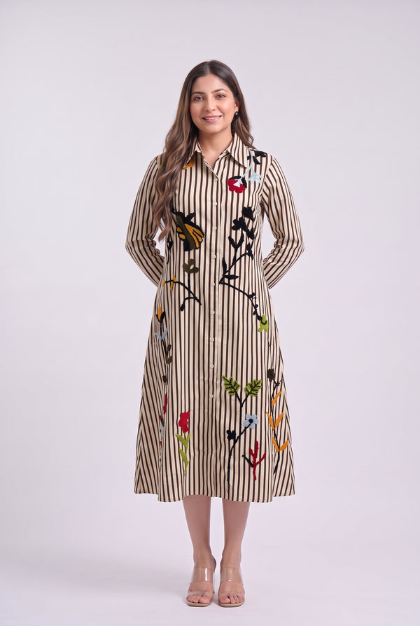 A woman modeling a long-sleeve midi shirt dress with vertical beige and brown stripes and colorful embroidered floral and butterfly motifs, set against a plain light gray studio background.