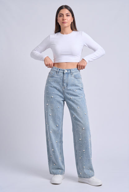 A woman modeling a pair of light-wash blue jeans with small white bead embellishments, paired with a white long-sleeve crop top and white sneakers, against a light gray studio background.