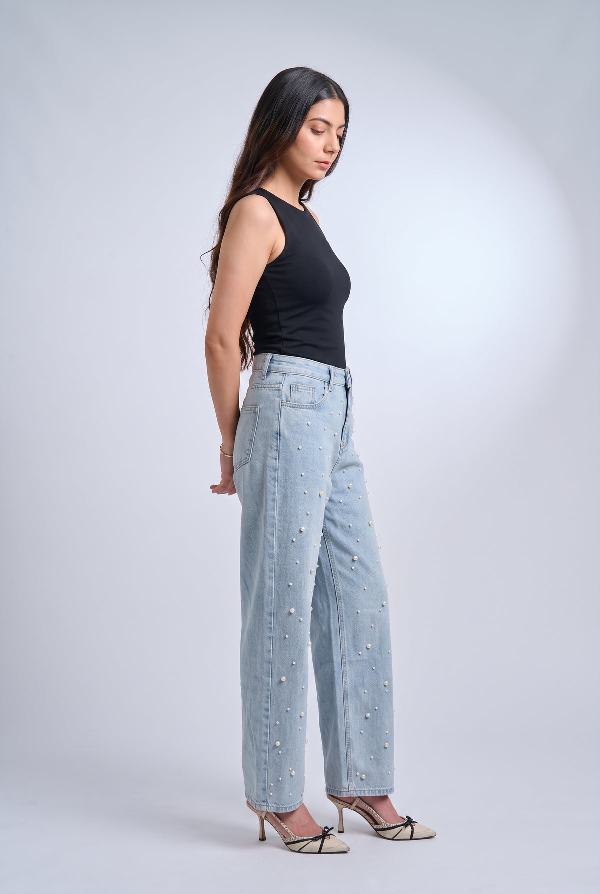 A woman stands in profile wearing light-wash wide-leg jeans with small pearl embellishments, a black sleeveless top, and beige pointed-toe heels against a light gray studio background.
