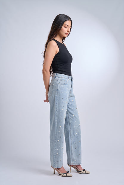 A woman stands in profile wearing light-wash wide-leg jeans with small pearl embellishments, a black sleeveless top, and beige pointed-toe heels against a light gray studio background.
