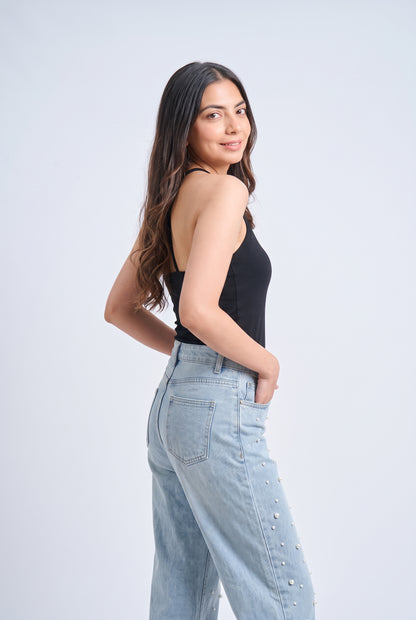 A woman posing in a studio wearing a black tank top and light wash blue jeans with silver stud embellishments on the side, set against a light gray background.