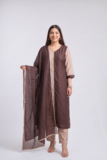 A woman modeling a traditional three-piece salwar kameez set in brown and beige, featuring a brown long kurta with beige side panels, matching beige trousers, and a brown dupatta with a beige border, shot against a light gray studio background.