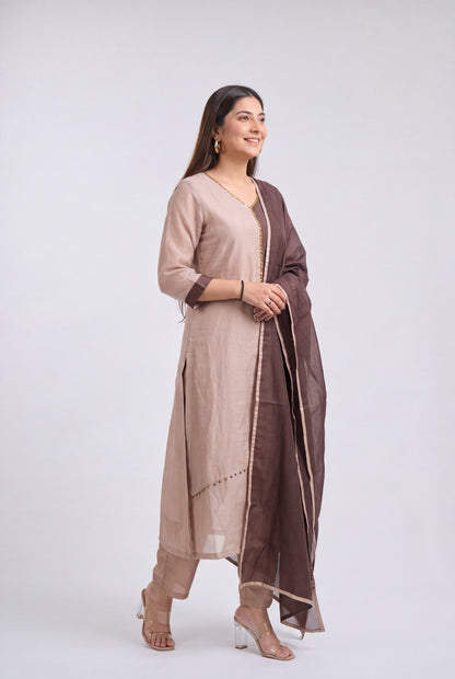 A woman modeling a traditional Indian outfit consisting of a beige kurta, matching trousers, and a dark brown dupatta with gold border details, set against a plain light gray background.