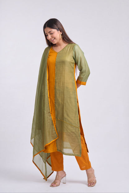 A woman modeling a traditional Indian outfit consisting of a mustard yellow kurta and pants with a sage green sheer dupatta, set against a plain light gray studio background.